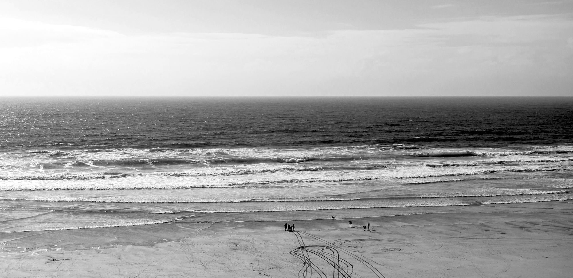 An empty Cornish beach from above: surf rolling in, a few walkers at the tideline, and tyre tracks looping across the sand
